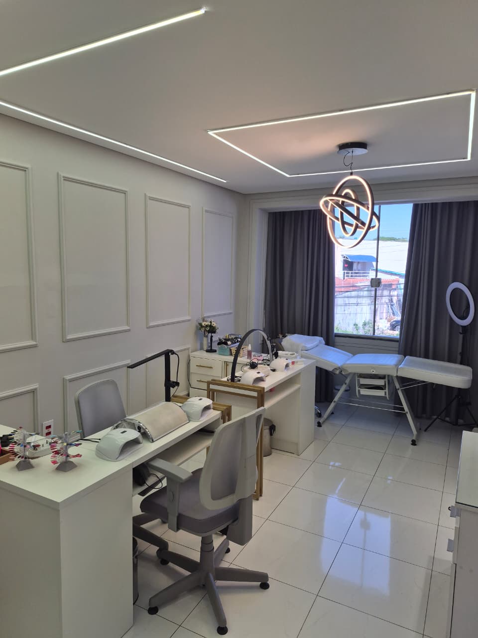 Sala com bancadas de unhas, maca junto à janela e iluminação no teto